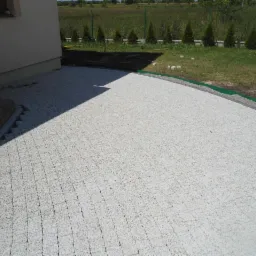 Jasnoszara kostka brukowa ułożona przy domu, widok z góry. Częściowo ułożona, z widocznym obrzeżem i fragmentem trawnika w tle. Słoneczny dzień.