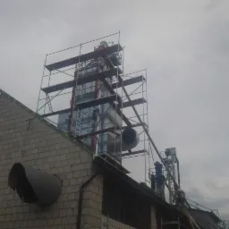 Montaż wentylacji na dachu budynku z szarej cegły, widoczne rusztowania i pracownicy. Duże rury wentylacyjne wystają z dachu, pochmurne niebo w tle.