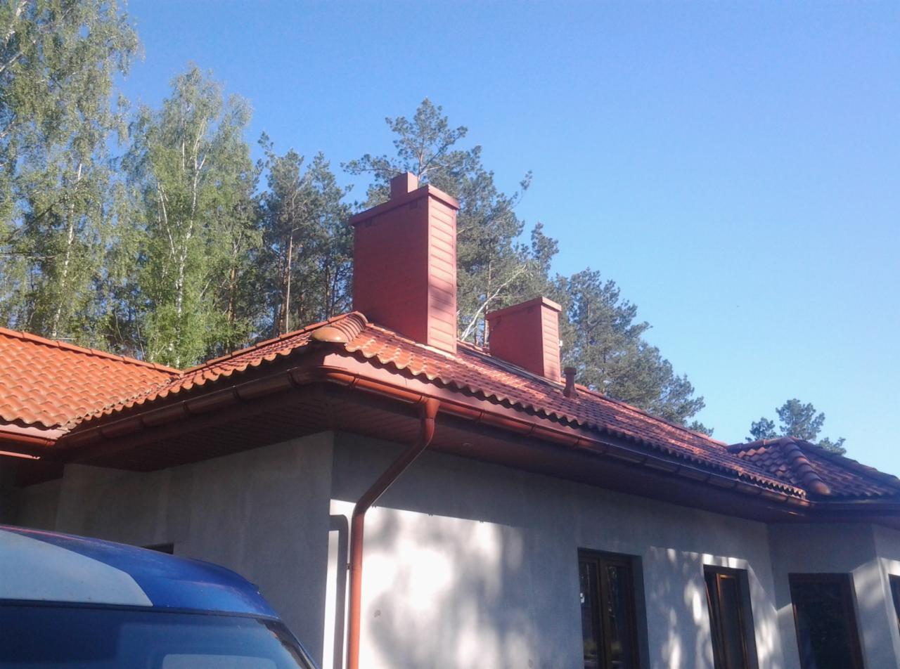 Czerwony dach z dachówki ceramicznej i dwa kominy na tle błękitnego nieba i zielonych drzew. Widoczne obróbki blacharskie i rynny w kolorze dachu.