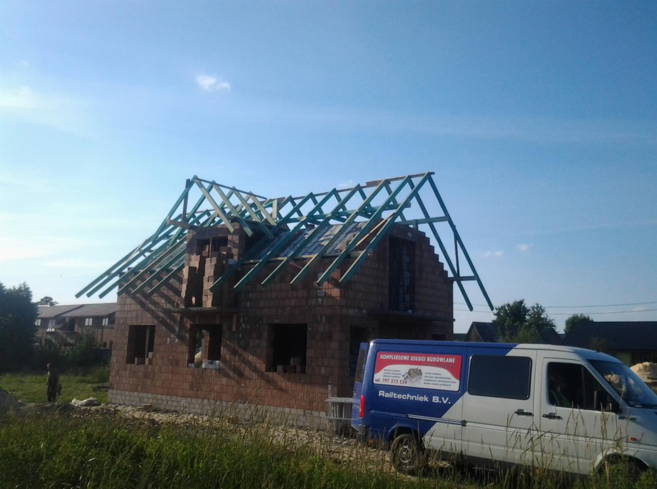 Surowy stan domu z cegły z częściowo wykonaną więźbą, na tle błękitnego nieba. Na pierwszym planie trawa i samochód dostawczy z reklamą firmy.