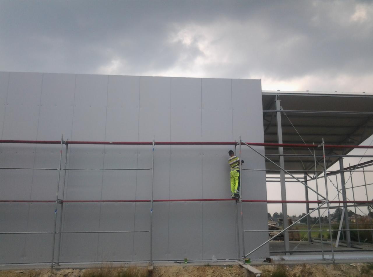 Pracownik w odblaskowej odzieży na rusztowaniu montuje panele ścienne na konstrukcji hali, widoczny fragment dachu i pochmurne niebo.