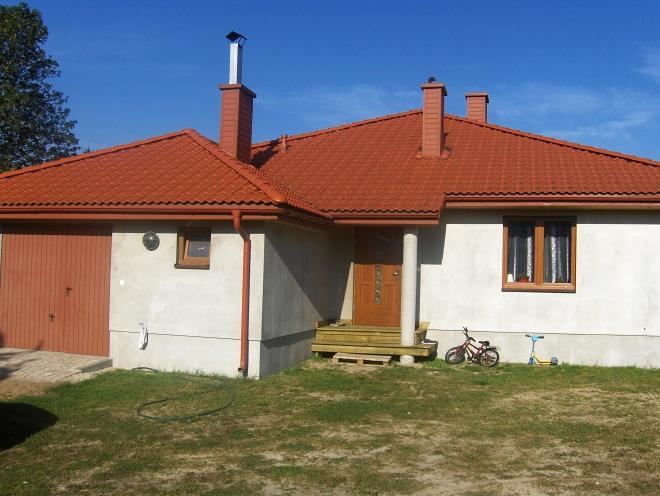 Dom jednorodzinny z czerwonym dachem, widoczne rynny i obróbki blacharskie. Na trawniku rower i hulajnoga dziecięca.