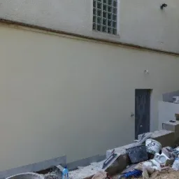 Otynkowana ściana zewnętrzna budynku z widocznymi materiałami budowlanymi i narzędziami na ziemi, fragment drabiny po lewej stronie.