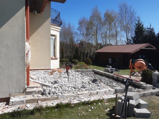 Demontaż starej wylewki fundamentowej przy budynku jednorodzinnym, widoczny gruz, młot pneumatyczny i betoniarka w ogrodzie.