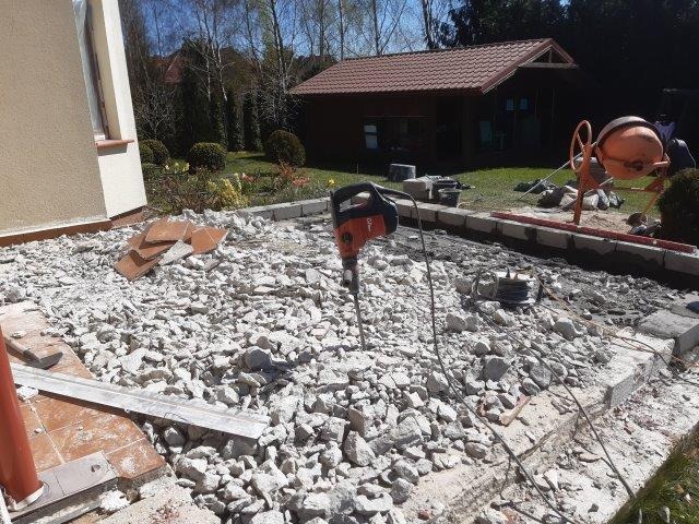 Teren przed domem w trakcie prac ziemnych: gruz, rozbita posadzka, młot pneumatyczny wbity w podłoże, betoniarka w tle, widoczne fundamenty.