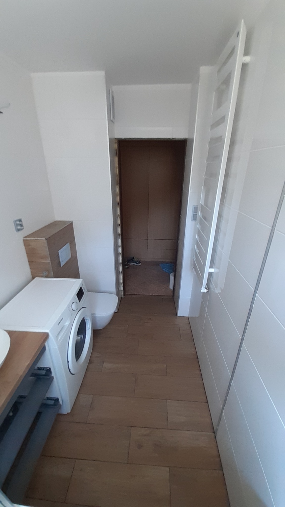 Wnętrze łazienki w trakcie prac wykończeniowych, widoczna pralka, toaleta, grzejnik drabinkowy i drewnopodobna podłoga, z otwartym przejściem do innego pomieszczenia, gdzie widać buty i niebieską...