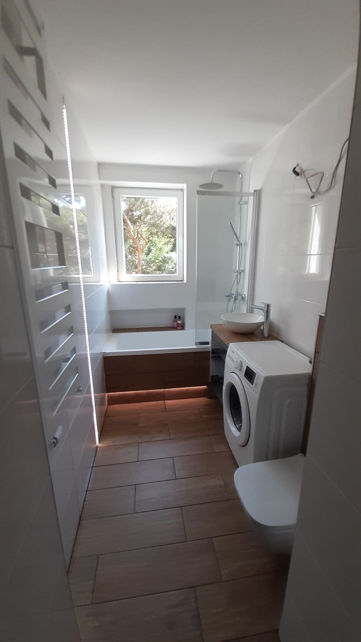 Nowoczesna łazienka z drewnopodobną podłogą, białą armaturą, pralką i oświetleniem LED pod wanną.