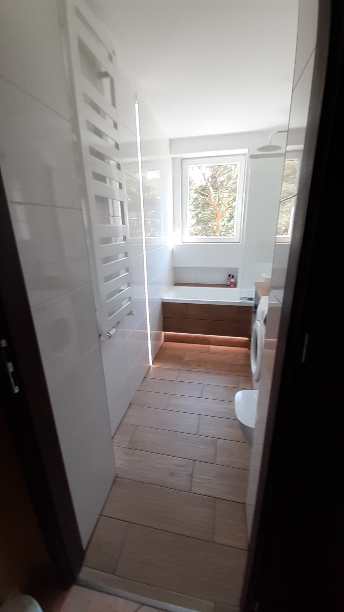 Wąska łazienka z drewnopodobną podłogą, białymi płytkami ściennymi, grzejnikiem drabinkowym, pralką i oknem z widokiem na drzewa.