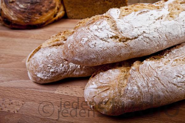 Trzy bagietki z mąką na drewnianym blacie, z widocznym pieczywem w tle. Ujęcie z góry podkreśla teksturę chleba i naturalne światło.