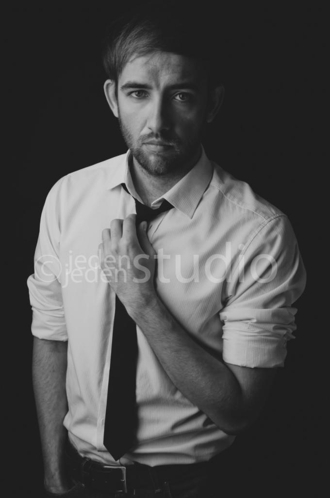 Czarno-biały portret mężczyzny w koszuli, poluzowującego krawat, na ciemnym tle. Ujęcie od pasa w górę, z delikatnym oświetleniem akcentującym rysy twarzy.