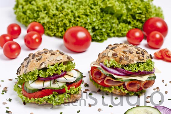 Dwa apetyczne, pełnoziarniste sandwicze z warzywami, szynką i serem na białym tle, otoczone świeżymi pomidorami i sałatą. Jasne, studyjne oświetlenie.