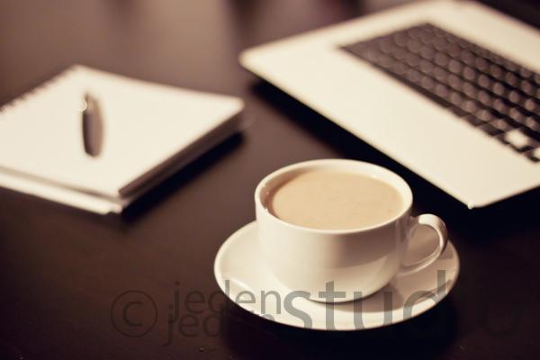 Kawa z mlekiem w białej filiżance na biurku obok otwartego laptopa i notatnika ze srebrnym długopisem. Ujęcie z bliska w ciepłej tonacji.