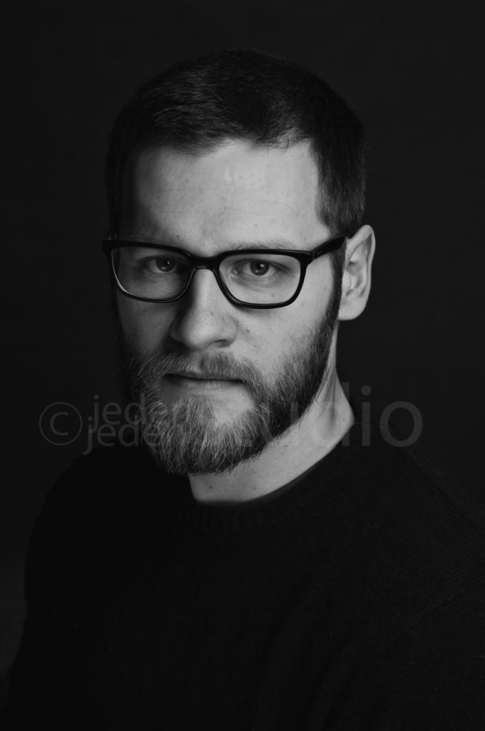 Czarno-biały portret brodatego mężczyzny w okularach na ciemnym tle, z widocznym logo fotografa w lewym dolnym rogu.