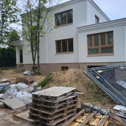 OL-BAU ŁUKASZ WĄWOŹNY - Biały, dwukondygnacyjny dom z brązowymi oknami w trakcie budowy, widoczne materiały budowlane i ogrodzenie tymczasowe na niezagospodarowanym terenie.