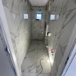 OL-BAU ŁUKASZ WĄWOŹNY - Wnętrze łazienki w trakcie wykańczania: białe płytki imitujące marmur na ścianach i podłodze, czerwone krzyżyki dystansowe, widoczne instalacje. Otwór drzwiowy z lewej.