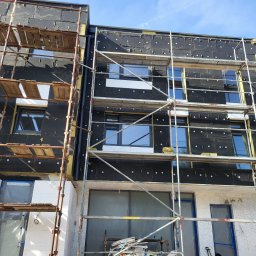 OL-BAU ŁUKASZ WĄWOŹNY - Budynek w trakcie ocieplania, widoczne rusztowania, czarna membrana i białe płyty izolacyjne. Częściowo otynkowana elewacja. Słoneczny dzień.