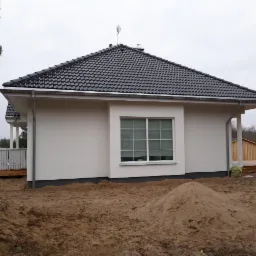 Jasny dom jednorodzinny z ciemną dachówką, świeżo po nałożeniu jasnego tynku, widoczna sterta piasku przed budynkiem.