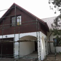 Dom jednorodzinny z drewnianą elewacją w trakcie malowania, widoczne rusztowania i fragmenty pomalowanej na biało dolnej części budynku.