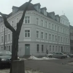 Odnowiona kamienica w pastelowych kolorach z szarym wykończeniem, widok z ulicy w pochmurny dzień, z zaparkowanymi samochodami i drzewami bez liści.