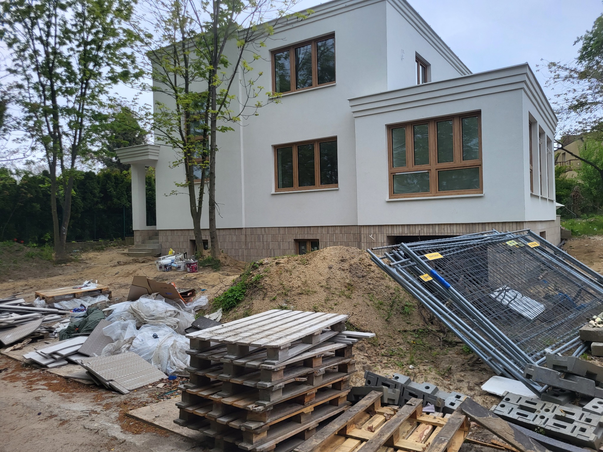 Biały, dwukondygnacyjny dom z brązowymi oknami w trakcie budowy, widoczne materiały budowlane i ogrodzenie tymczasowe na niezagospodarowanym terenie.