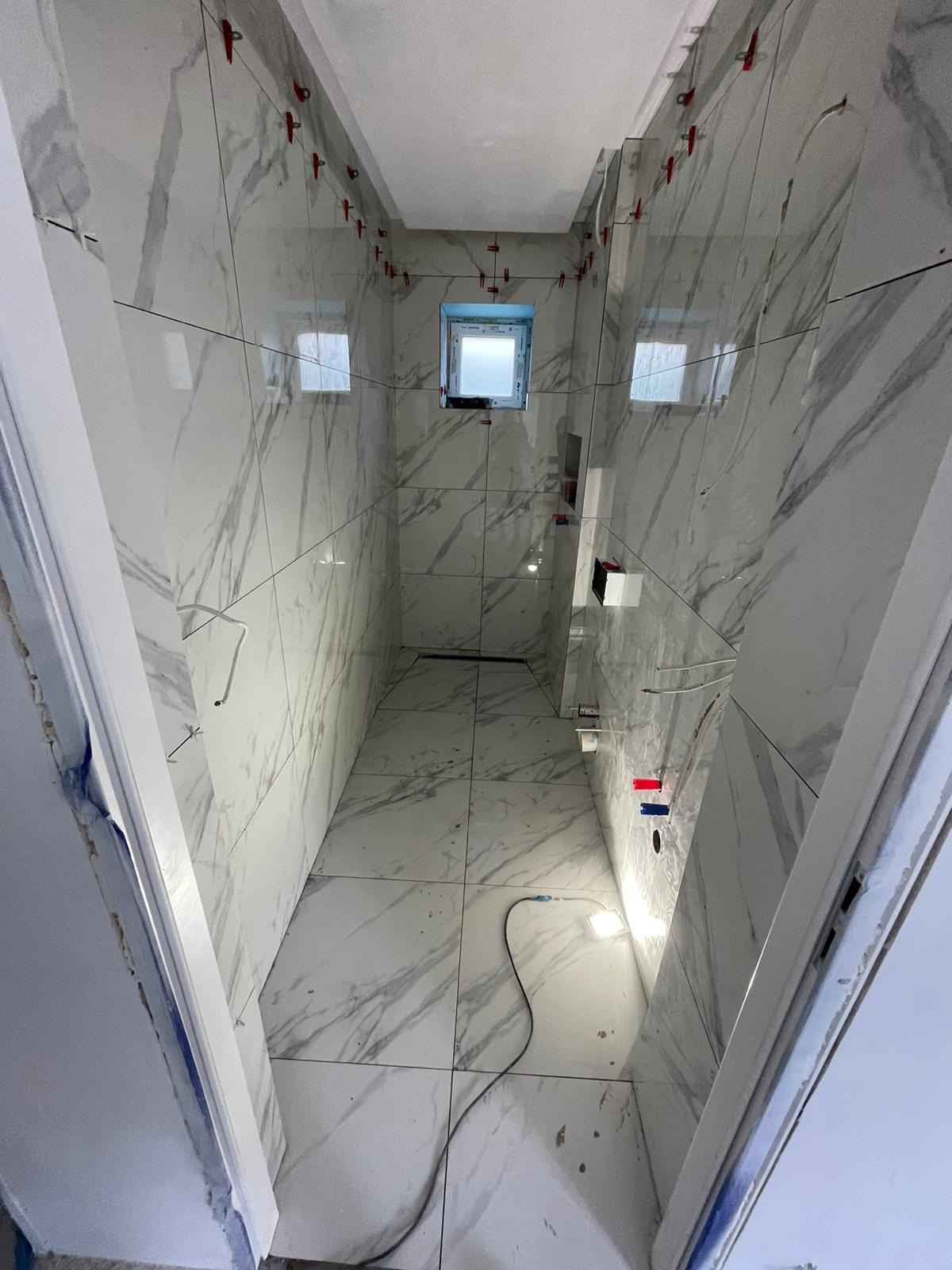 Wnętrze łazienki w trakcie wykańczania: białe płytki imitujące marmur na ścianach i podłodze, czerwone krzyżyki dystansowe, widoczne instalacje. Otwór drzwiowy z lewej.
