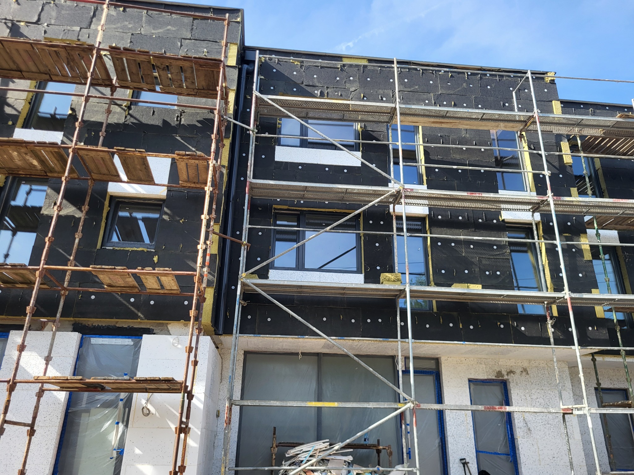 Budynek w trakcie ocieplania, widoczne rusztowania, czarna membrana i białe płyty izolacyjne. Częściowo otynkowana elewacja. Słoneczny dzień.