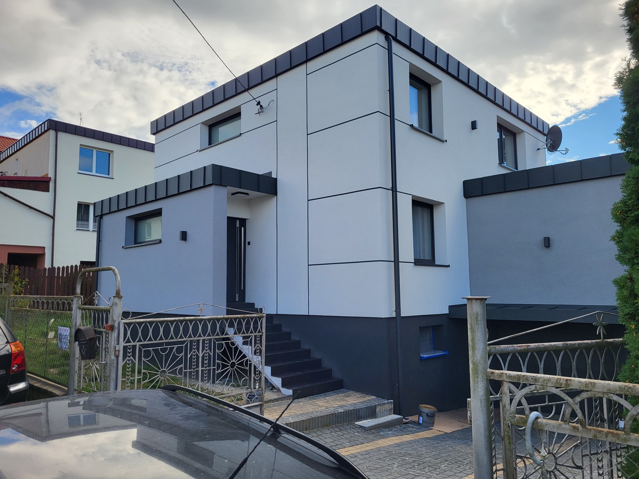 Dwukondygnacyjny dom z elewacją w odcieniach szarości i bieli, z czarnymi akcentami. Widoczne schody wejściowe, metalowe ogrodzenie i fragment dachu samochodu na pierwszym planie.