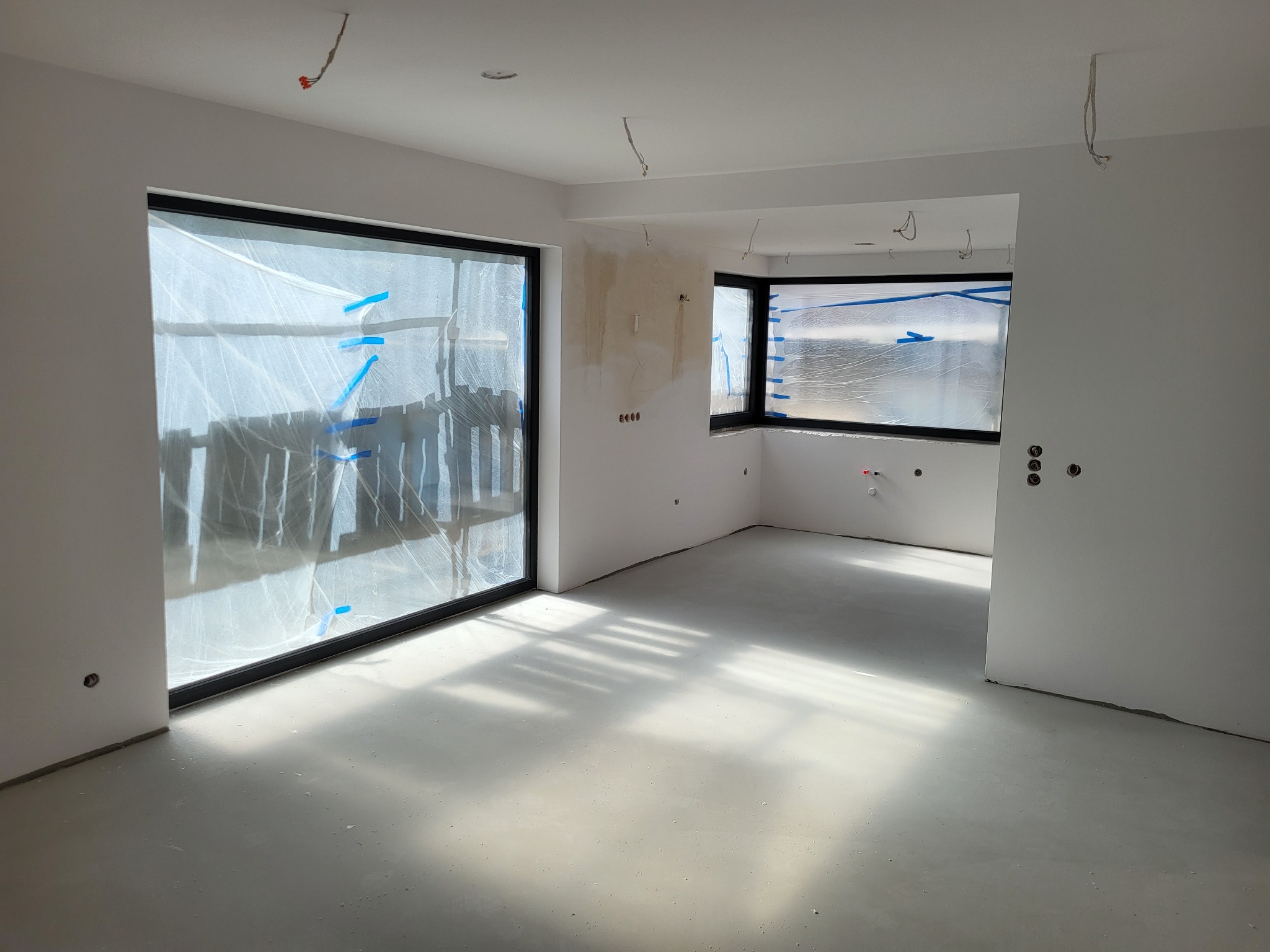 Wnętrze w trakcie prac wykończeniowych: świeżo wylana posadzka, okna zabezpieczone folią malarską, ściany przygotowane do malowania, widoczne kable wystające z sufitu.