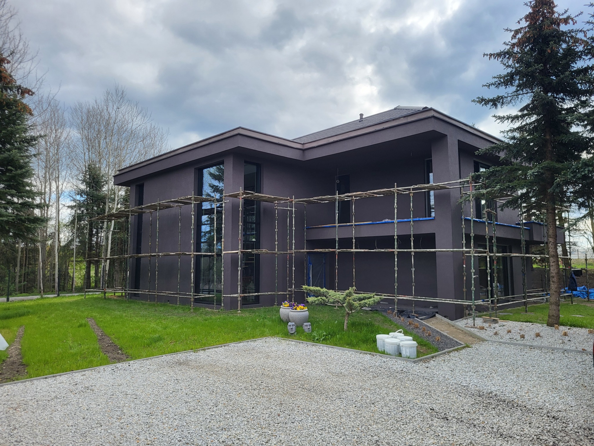 Nowoczesny dom z ciemną elewacją w trakcie prac wykończeniowych, otoczony rusztowaniami i zielenią. Widoczne pojemniki z farbą i dekoracje ogrodowe.