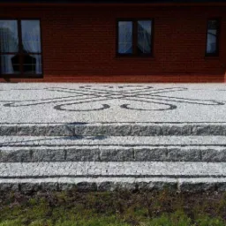 Granitowe schody prowadzą na taras z kostki granitowej z dekoracyjnym wzorem z ciemniejszego kamienia, na tle ceglanego budynku z oknami.