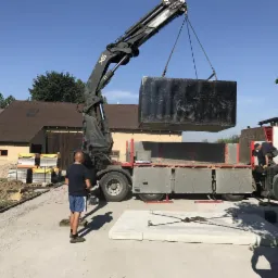 Montaż masywnego, czarnego bloku kamiennego przy użyciu dźwigu samochodowego na placu budowy, widoczny pracownik na skrzyni ładunkowej ciężarówki i przygotowany betonowy fundament.