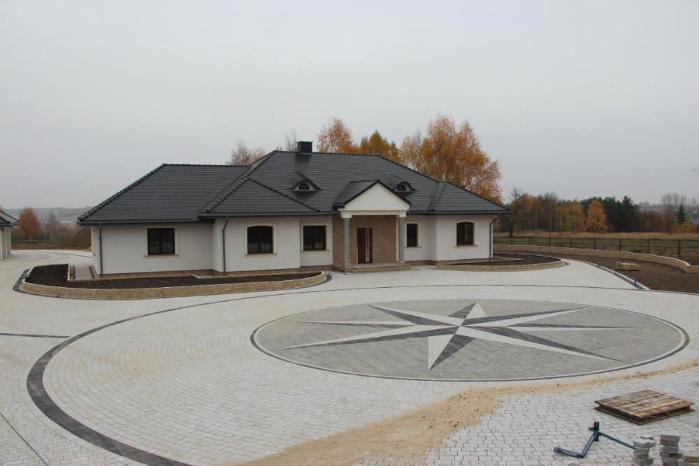 Rozległy podjazd z kostki brukowej w kształcie rozety kompasu, otaczający nowoczesny dom z ciemnym dachem i jasną elewacją, widok z perspektywy ogólnej w pochmurny dzień.