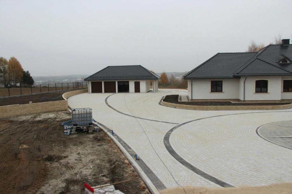 Szeroki, zakrzywiony podjazd wyłożony jasnoszarą kostką brukową z ciemnoszarymi akcentami, prowadzący do domu i garażu, z widocznym zbiornikiem na wodę i materiałami budowlanymi na obrzeżach.