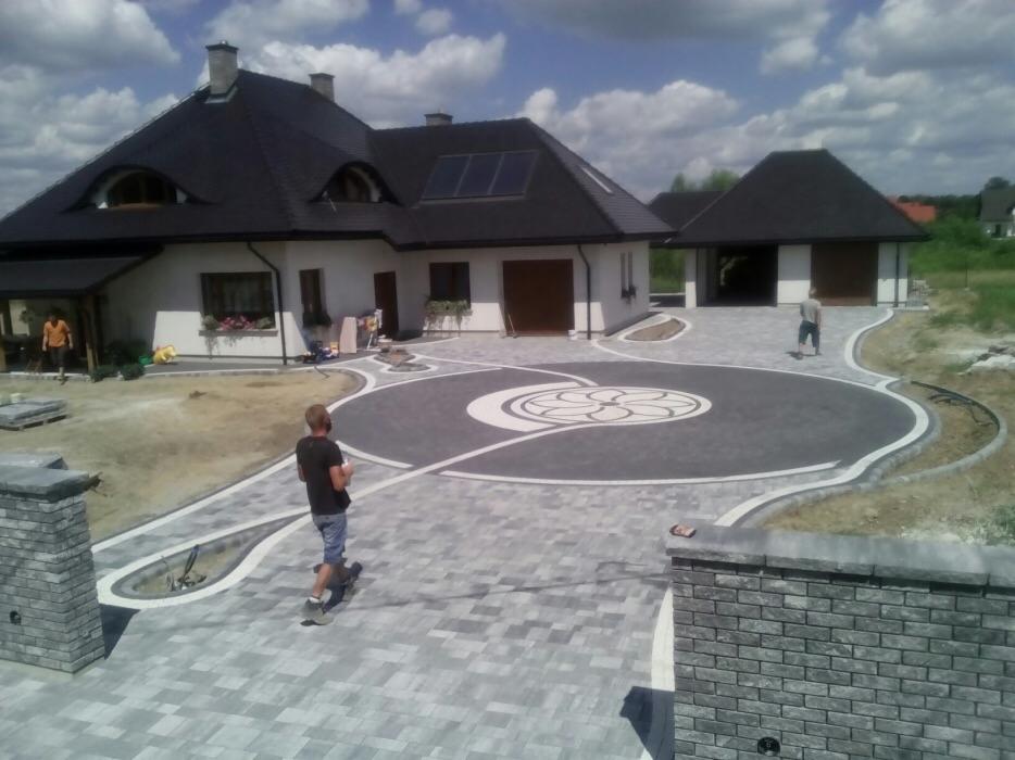Szeroki podjazd z szarej kostki brukowej z dekoracyjnym wzorem w kształcie kwiatu na okrągłym placu przed domem z ciemnym dachem i garażem.
