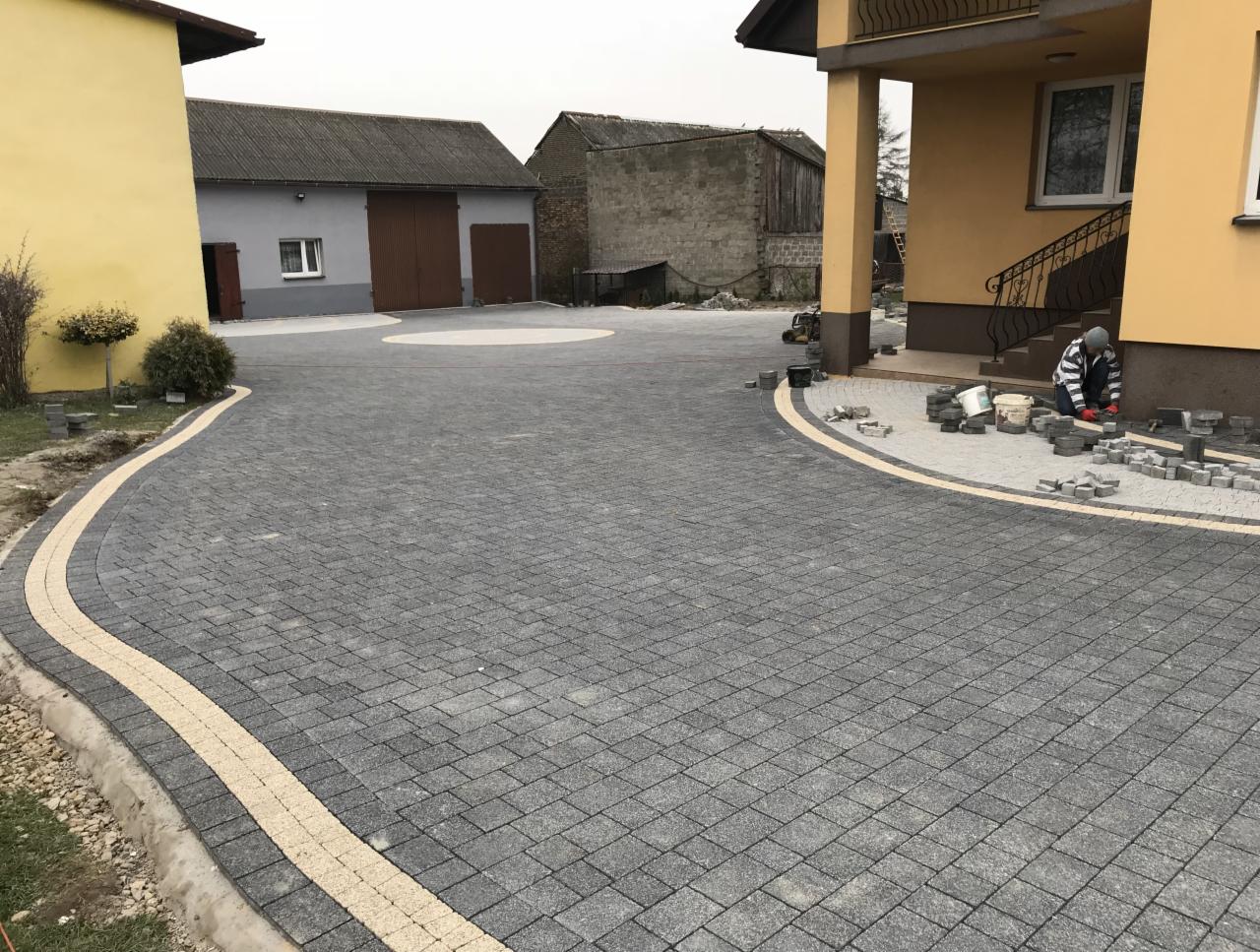 Układanie szarej kostki brukowej z jasnym obramowaniem na podjeździe, widoczny pracownik w paski układający kostkę przy schodach domu.