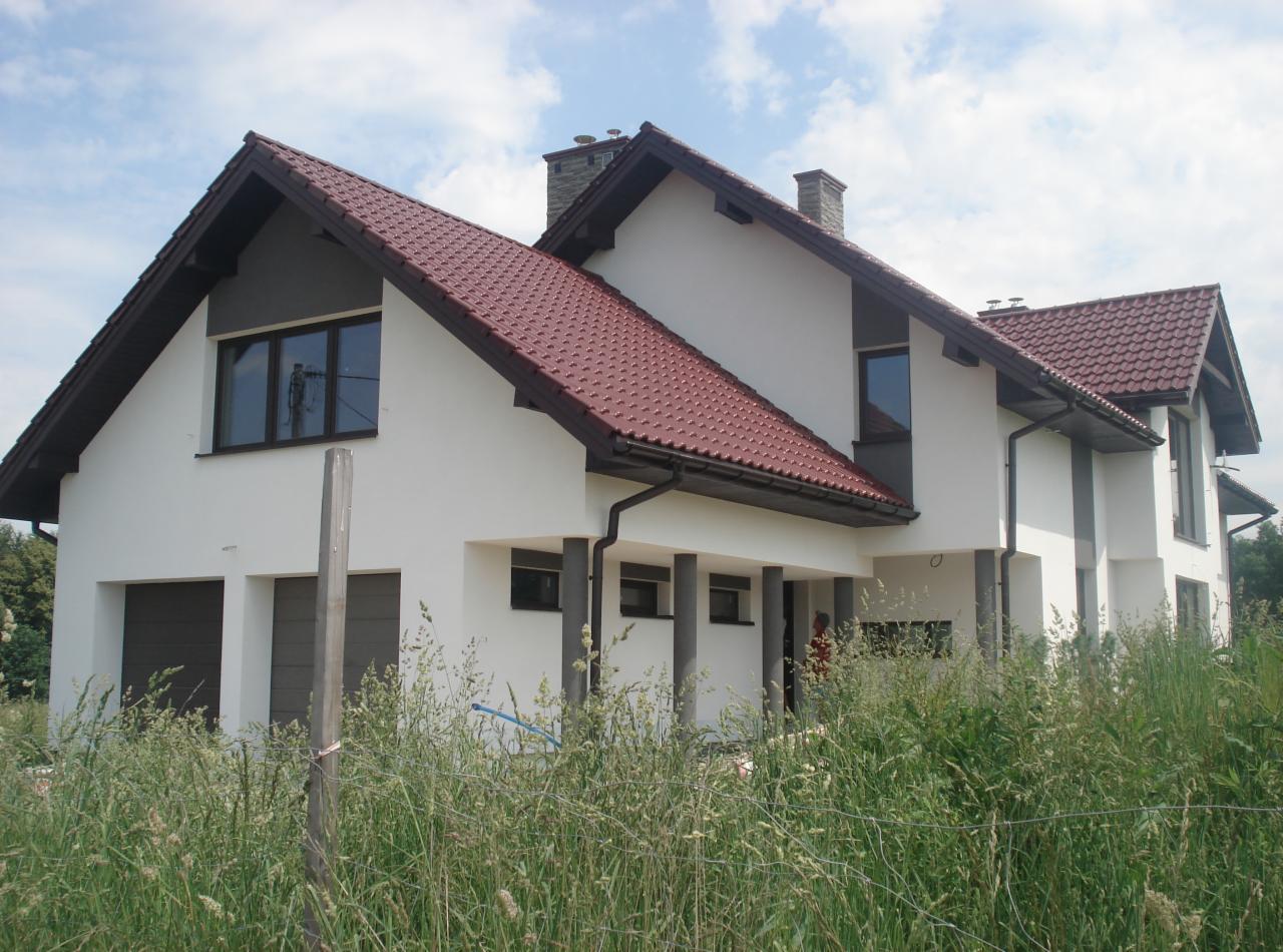 Nowoczesny dom z białą elewacją i czerwoną dachówką, otoczony wysoką trawą. Widoczne szare elementy wykończeniowe i brązowe okna.
