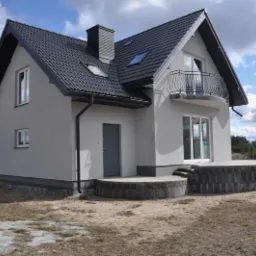 Nowoczesny dom jednorodzinny z balkonem i ciemną dachówką, szara elewacja, widok z zewnątrz na tle błękitnego nieba z chmurami.