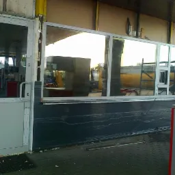 Szklana ściana z oknem i drzwi PCV w szarych ramach, zamontowane w budynku stacji benzynowej, widoczne elementy konstrukcyjne i otoczenie zewnętrzne.