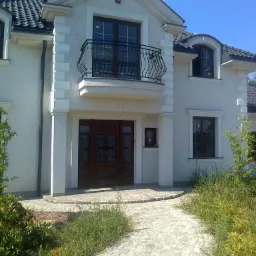 Elewacja domu jednorodzinnego z drewnianymi oknami i kutą balustradą balkonową, widok na ozdobne drzwi wejściowe z kamienną ścieżką.