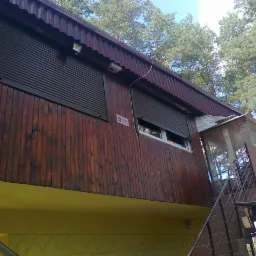 Drewniany dom z brązowymi roletami zewnętrznymi na oknach, widoczne schody wejściowe i fragmenty drzew w tle.