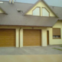 Dom z dwoma brązowymi bramami garażowymi i oknami z brązowymi roletami zewnętrznymi, widziany z bliska w pochmurny dzień.