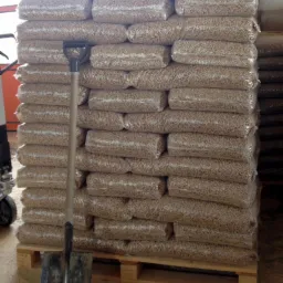 Stos worków z pelletem na palecie, szufla oparta o worki. Widoczny fragment wózka paletowego. Magazyn z zapasem pelletu.