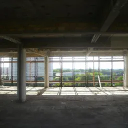 Surowy stan wnętrza budynku w trakcie budowy. Widoczne kolumny, strop, niezabudowane otwory okienne z widokiem na zieleń i budynki w tle.