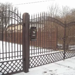 Kuta brama wjazdowa w kolorze ciemnego brązu z ozdobnymi elementami i skrzynką na listy, widoczna na tle drewnianego ogrodzenia i zaśnieżonego podjazdu.