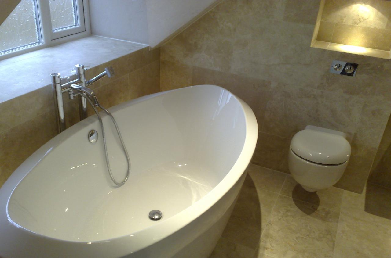 Nowoczesna łazienka z wolnostojącą białą wanną, chromowaną armaturą i podwieszaną toaletą na tle beżowych marmurowych płytek i wnęki z oświetleniem.