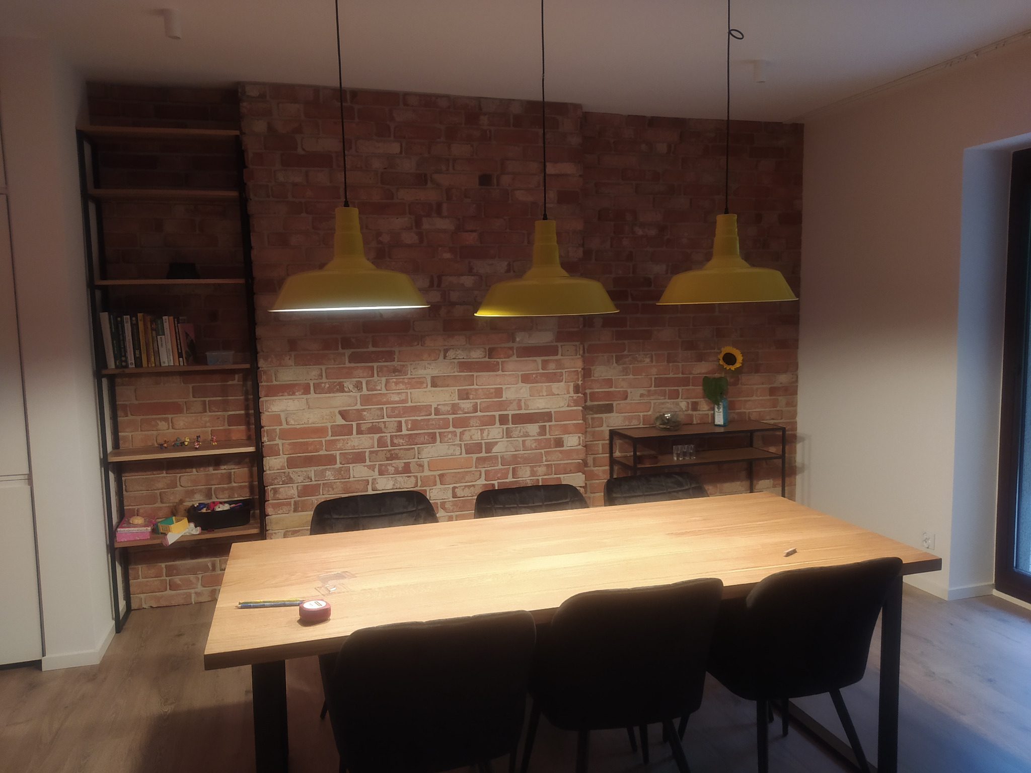 Jadalnia z ceglaną ścianą, drewnianym stołem i trzema żółtymi lampami wiszącymi; na stole leży miarka i ołówek; w tle regał z książkami i dekoracjami.