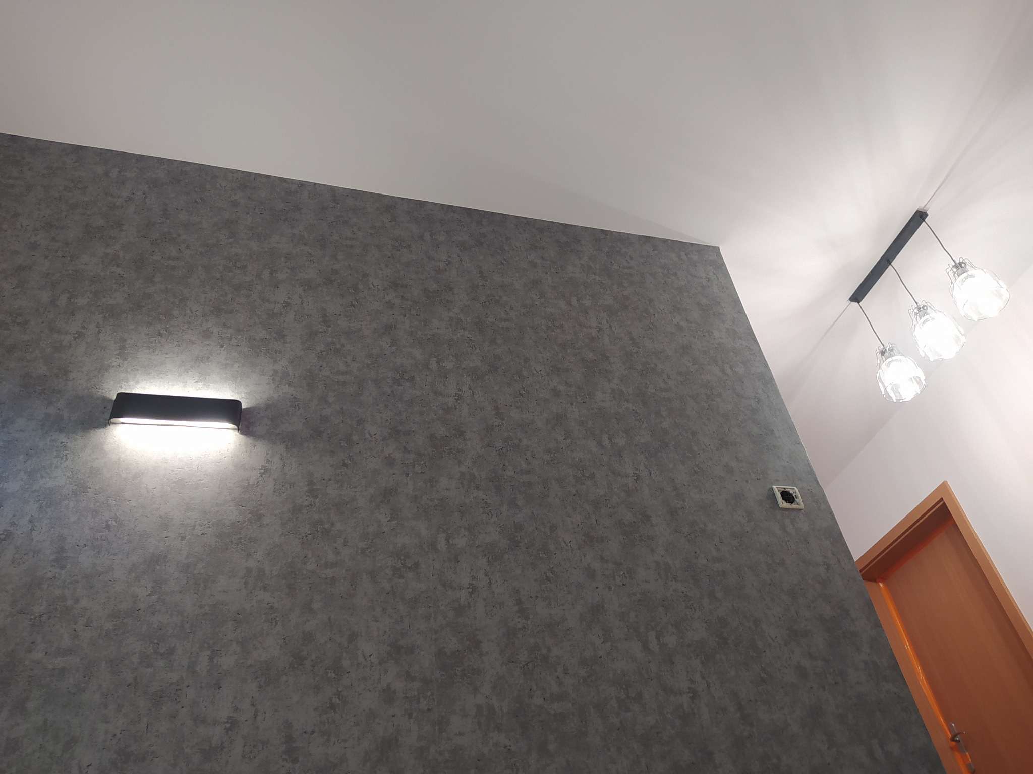 Szara ściana z fakturą, oświetlona kinkietem i lampą sufitową, widoczny fragment drzwi i gniazdko elektryczne bez osłonki.