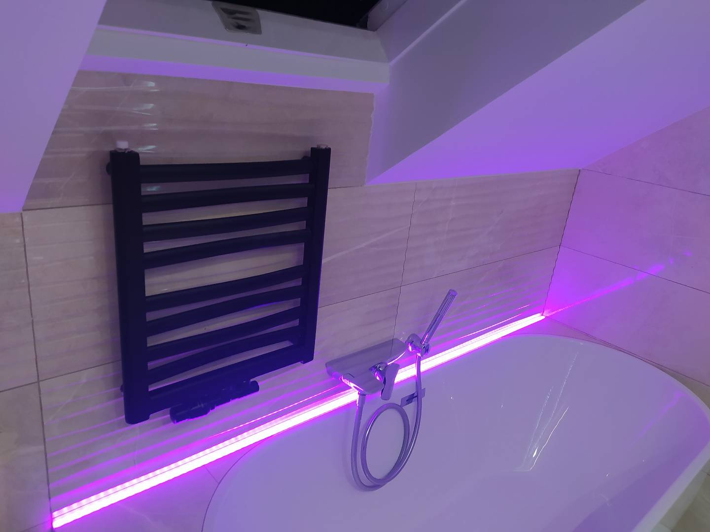 Łazienka na poddaszu z wanną, chromowaną baterią i czarnym grzejnikiem drabinkowym, oświetlona fioletowym światłem LED.