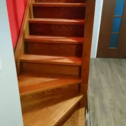 Wnętrze domu z nowymi, drewnianymi schodami o ciepłym odcieniu, prowadzącymi w górę, z widoczną czerwoną ścianą po lewej stronie i fragmentem podłogi imitującej drewno.