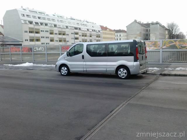 Srebrny van Opel Vivaro zaparkowany na ulicy miejskiej, widok z boku, na tle budynku mieszkalnego i metalowego ogrodzenia z graffiti.
